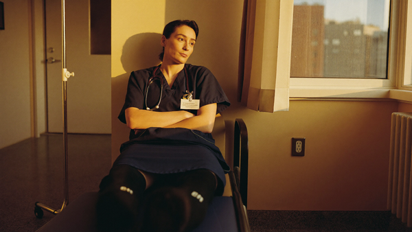 Nurse in scrubs resting on a hospital corridor bench at the end of a long shift, wearing black knee-high compression socks