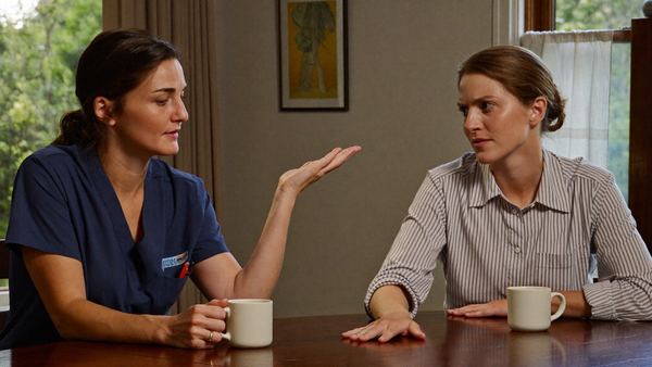 Two sisters talking over coffee at a kitchen table, one in navy scrubs telling a story, the other listening skeptically